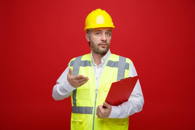 Hombre constructor en uniforme de construcción y casco de seguridad sosteniendo portapapeles mirando confundido levantando el brazo con disgusto de pie sobre fondo rojo.