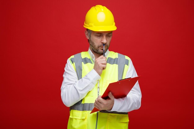 Hombre constructor en uniforme de construcción y casco de seguridad sosteniendo portapapeles mirando desconcertado pensando de pie sobre fondo rojo.