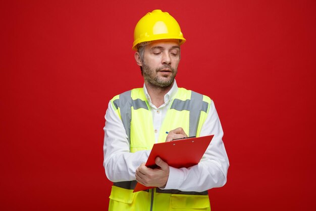 Hombre constructor en uniforme de construcción y casco de seguridad sosteniendo portapapeles mirándolo con cara seria haciendo notas de pie sobre fondo rojo.