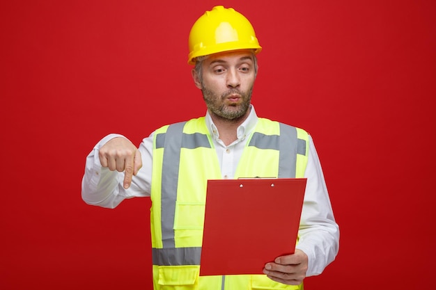 Hombre constructor en uniforme de construcción y casco de seguridad sosteniendo portapapeles mirándolo confundido apuntando con el dedo índice hacia abajo de pie sobre fondo rojo