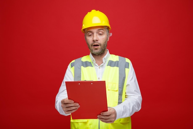 Hombre constructor en uniforme de construcción y casco de seguridad sosteniendo portapapeles mirándolo sorprendido de pie sobre fondo rojo