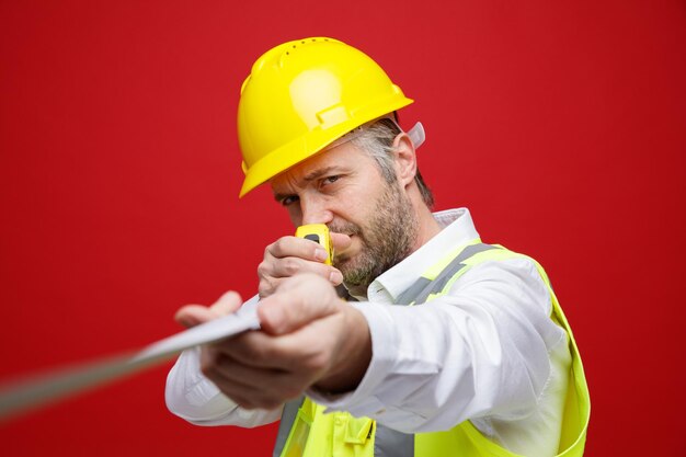 Hombre constructor en uniforme de construcción y casco de seguridad sosteniendo una regla apuntándola a la cámara mirando confiado de pie sobre fondo rojo.