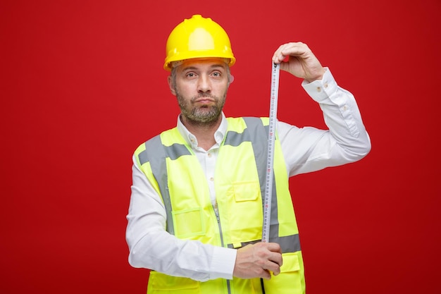 Hombre constructor en uniforme de construcción y casco de seguridad sosteniendo una regla mirando a la cámara con cara seria de pie sobre fondo rojo.