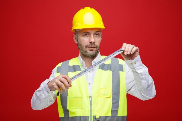 Hombre constructor en uniforme de construcción y casco de seguridad sosteniendo una regla mirando a la cámara con expresión segura de pie sobre fondo rojo.