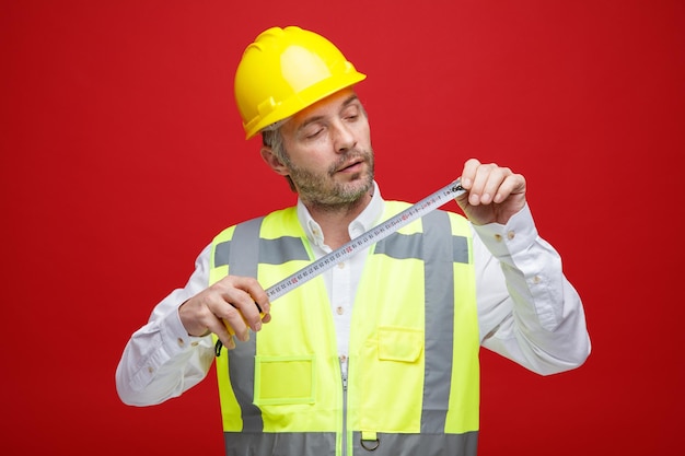 Hombre constructor en uniforme de construcción y casco de seguridad sosteniendo una regla mirándolo intrigado de pie sobre fondo rojo.