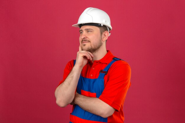 Hombre constructor con uniforme de construcción y casco de seguridad tocando su mejilla y pensando en una mirada pensativa de pie sobre una pared rosa aislada