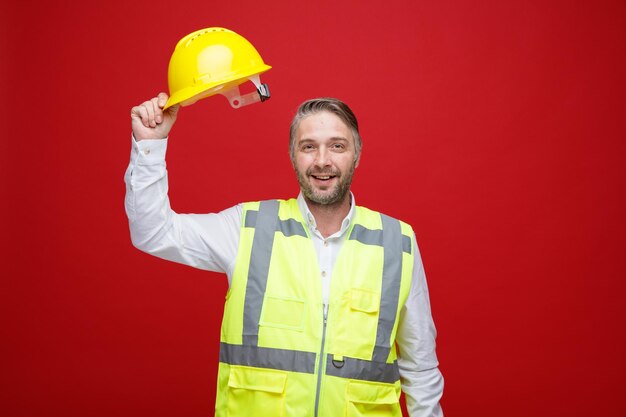 Hombre constructor en uniforme de construcción sosteniendo su casco de seguridad mirando a la cámara feliz y alegre sonriendo alegremente de pie sobre fondo rojo.