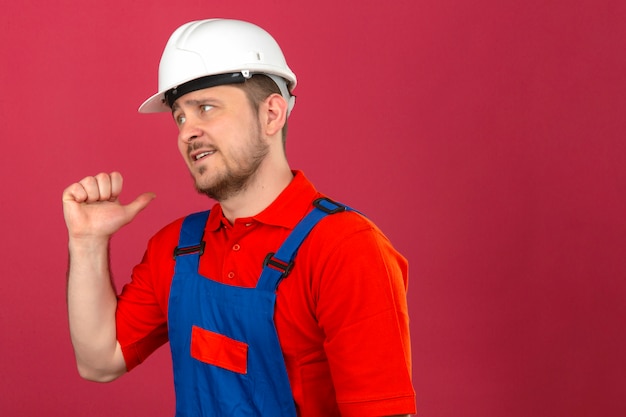 Hombre constructor vistiendo uniforme de construcción y casco de seguridad apuntando hacia atrás con la mano y el pulgar arriba disgustado de pie sobre la pared rosa oscuro aislada