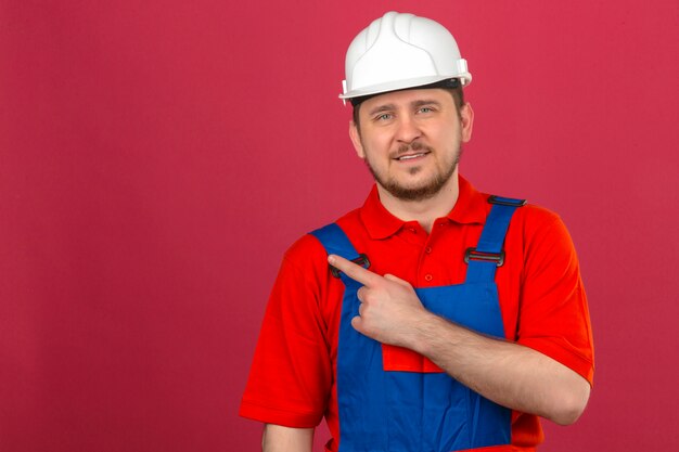 Hombre constructor vistiendo uniforme de construcción y casco de seguridad apuntando con el dedo para copiar el espacio mirando seguro de pie sobre la pared rosada aislada