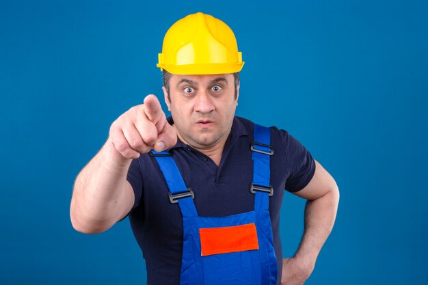 Hombre constructor vistiendo uniforme de construcción y casco de seguridad apuntando disgustado y frustrado a la cámara enojado y furioso contigo sobre la pared azul aislada