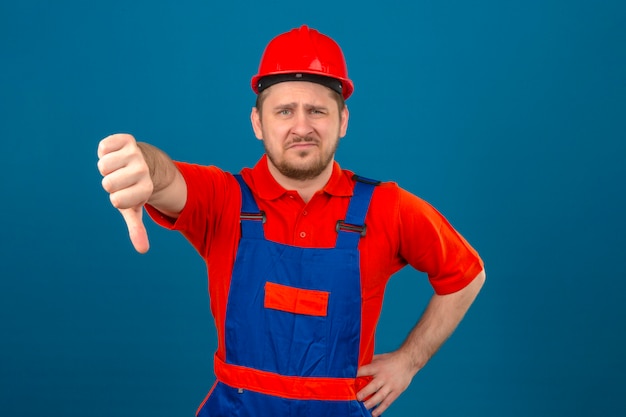 Hombre constructor vistiendo uniforme de construcción y casco de seguridad disgustado mostrando el pulgar hacia abajo de pie sobre la pared azul aislada