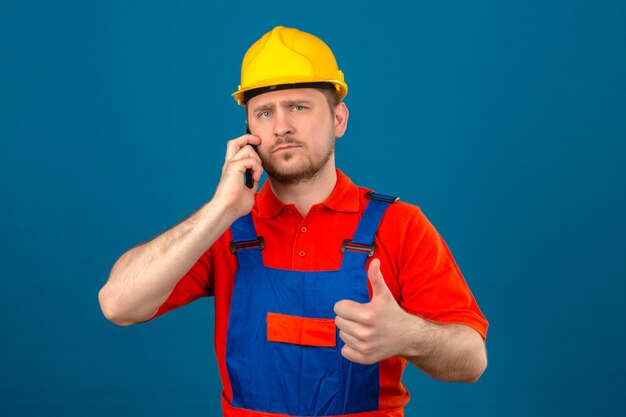Hombre constructor vistiendo uniforme de construcción y casco de seguridad hablando por teléfono móvil con cara seria mostrando el pulgar hacia arriba de pie sobre la pared azul aislada