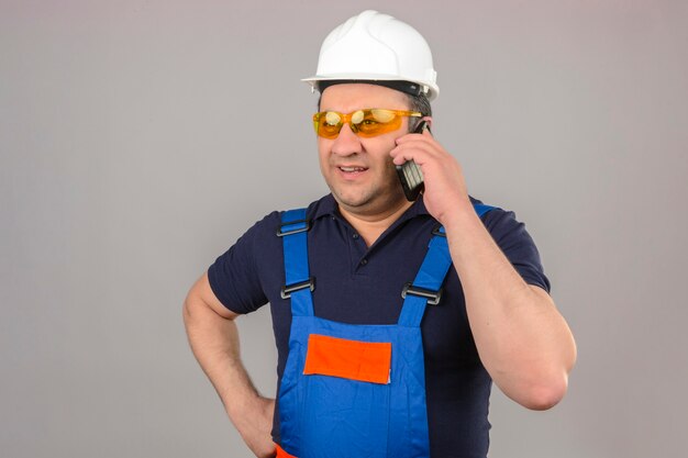 Hombre constructor vistiendo uniforme de construcción y casco de seguridad hablando por teléfono móvil sonriendo sobre pared blanca aislada