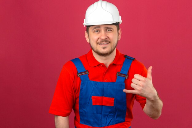 Hombre constructor vistiendo uniforme de construcción y casco de seguridad haciendo llamarme gesto mirando seguro de pie sobre pared rosa aislado