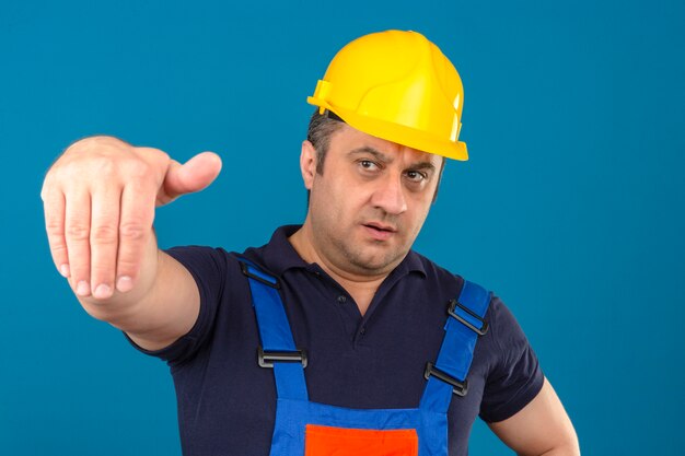 Hombre constructor vistiendo uniforme de construcción y casco de seguridad haciendo venir aquí gesto con la mano con cara seria sobre pared azul aislado