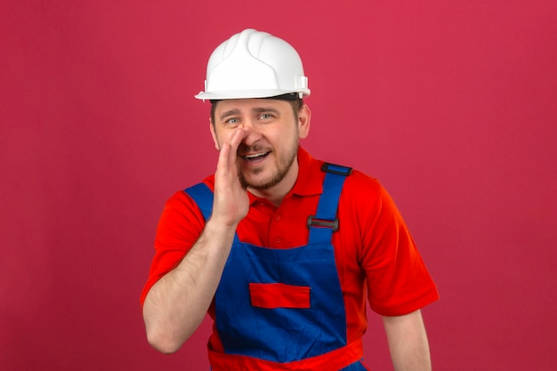 Hombre constructor vistiendo uniforme de construcción y casco de seguridad con una mano cerca de la boca contando un secreto de pie sobre la pared rosa oscuro aislada