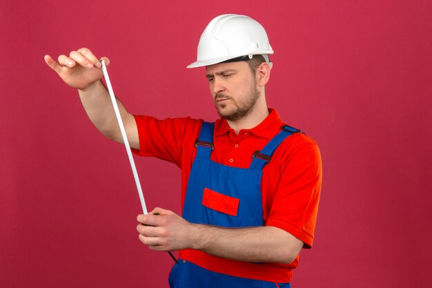 Hombre constructor vistiendo uniforme de construcción y casco de seguridad mirando cinta métrica en manos con cara seria de pie sobre pared rosa aislado