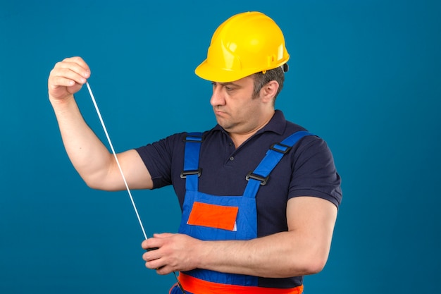Hombre constructor vistiendo uniforme de construcción y casco de seguridad mirando en cinta métrica en sus manos mirada pensativa sobre pared azul aislado