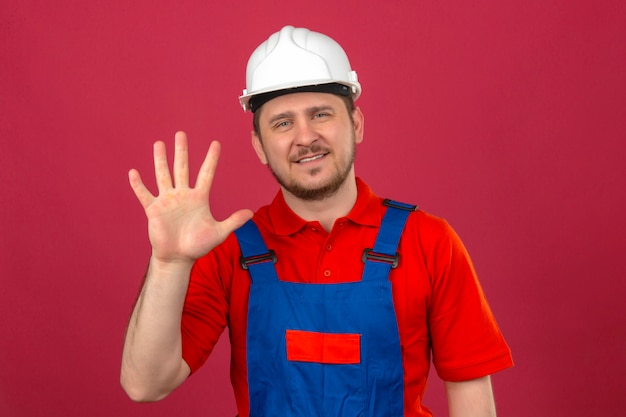 Hombre constructor vistiendo uniforme de construcción y casco de seguridad mostrando y apuntando hacia arriba con los dedos número cinco sonriendo confiados sobre pared rosa aislado