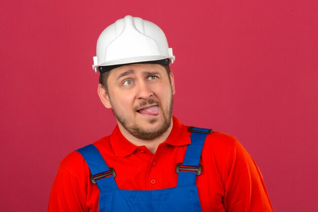 Hombre constructor vistiendo uniforme de construcción y casco de seguridad mostrando cara de mueca cruzando los ojos sacando la lengua sobre la pared rosada aislada