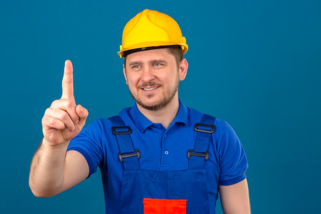 Foto gratuita hombre constructor vistiendo uniforme de construcción y casco de seguridad mostrando el número uno con el dedo mirando a un lado sonriendo amigable de pie sobre la pared azul aislada