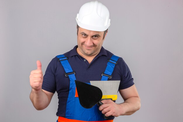 Hombre constructor vistiendo uniforme de construcción y casco de seguridad de pie con llana y espátula mostrando los pulgares con cara feliz y sonriendo sobre pared blanca aislada