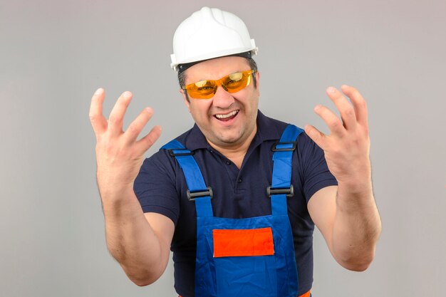 Hombre constructor vistiendo uniforme de construcción y casco de seguridad de pie loco y loco con expresión agresiva y los brazos levantados sobre la pared blanca aislada