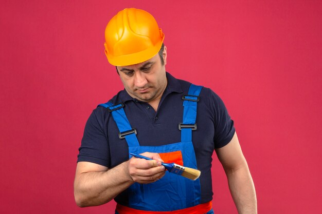 Hombre constructor vistiendo uniforme de construcción y casco de seguridad de pie con pincel y mirándolo con cara seria sobre pared rosa aislado
