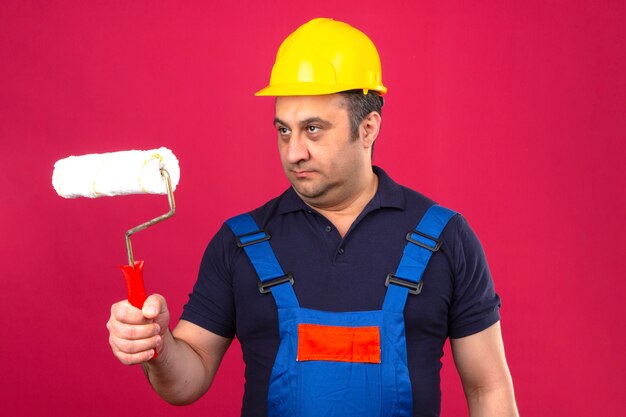 Hombre constructor vistiendo uniforme de construcción y casco de seguridad de pie con rodillo de pintura mirando a otro lado sobre pared rosa aislado