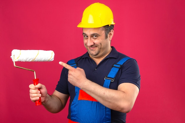 Foto gratuita hombre constructor vistiendo uniforme de construcción y casco de seguridad de pie con rodillo de pintura sonriendo y señalando con el dedo al rodillo de pintura sobre pared rosa aislado