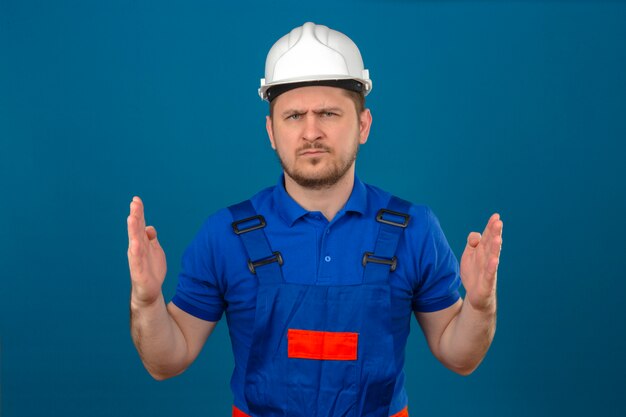 Hombre constructor vistiendo uniforme de construcción y casco de seguridad que muestra una señal de gran tamaño con el ceño fruncido símbolo de medida de pie sobre la pared azul aislada