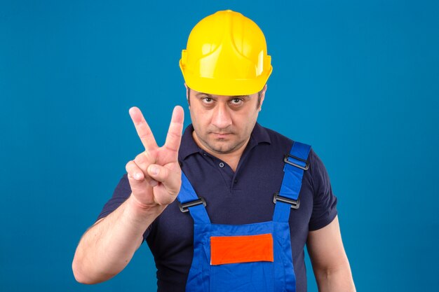 Hombre constructor vistiendo uniforme de construcción y casco de seguridad y señalando con el dedo como intimidación sobre pared azul aislada