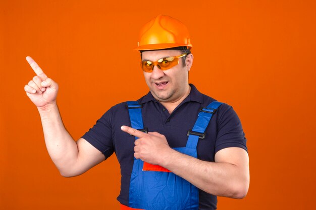 Hombre constructor vistiendo uniforme de construcción y casco de seguridad sonriendo con cara feliz y apuntando hacia el lado con los dedos sobre la pared naranja aislada