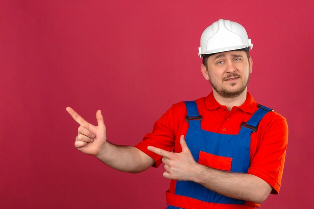 Hombre constructor vistiendo uniforme de construcción y casco de seguridad sonriendo y mirando a la cámara apuntando con las dos manos y los dedos al lado parado sobre la pared rosa aislada