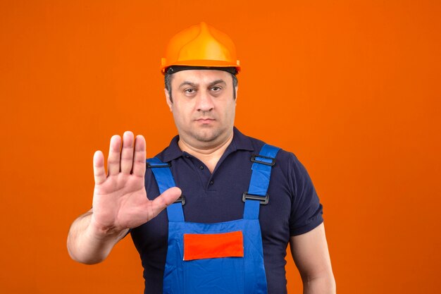 Hombre constructor vistiendo uniforme de construcción y casco de seguridad sosteniendo su mano haciendo gesto de parada sobre la pared naranja aislada