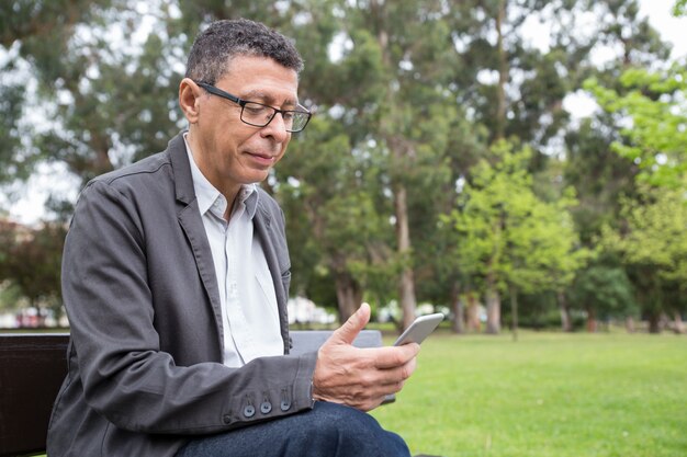 Hombre contento que usa teléfono inteligente y sentado en el banco en el parque