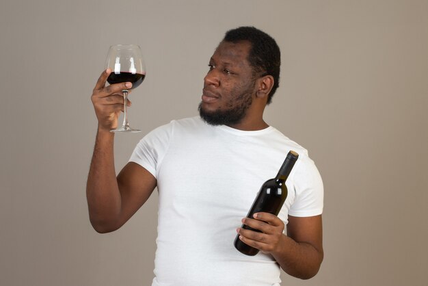 Hombre con copa de vino en una mano y botella de vino en la otra, de pie frente a la pared gris.