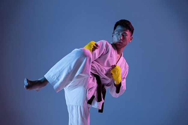 Hombre coreano confiado en kimono practicando combate cuerpo a cuerpo, artes marciales. Joven luchador con cinturón negro entrenando en pared degradada con luz de neón. Concepto de estilo de vida saludable, deporte.