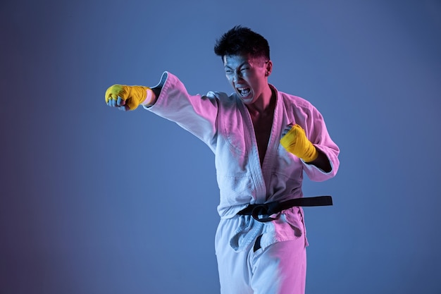 Foto gratuita hombre coreano confiado en kimono practicando combate cuerpo a cuerpo, artes marciales. joven luchador con cinturón negro entrenando en pared degradada con luz de neón. concepto de estilo de vida saludable, deporte.