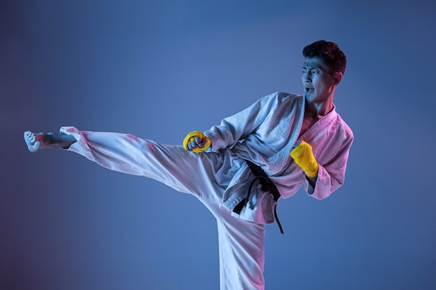 Hombre coreano confiado en kimono practicando combate cuerpo a cuerpo, artes marciales. Joven luchador con cinturón negro entrenando sobre fondo degradado en luz de neón. Concepto de estilo de vida saludable, deporte.