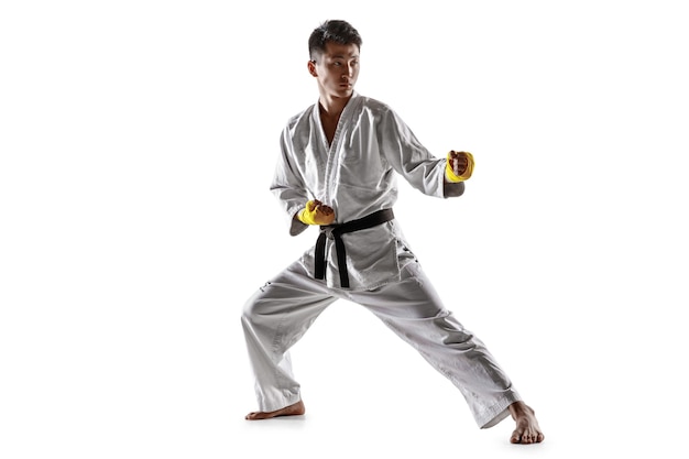 Hombre coreano confiado en kimono practicando combate cuerpo a cuerpo, artes marciales. Luchador masculino joven con entrenamiento de cinturón negro aislado en la pared blanca. Concepto de estilo de vida saludable, deporte.