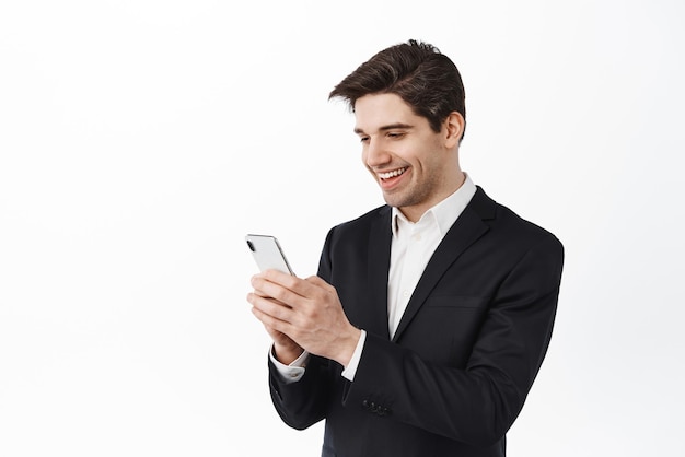 Hombre corporativo guapo chateando por teléfono y mirando la pantalla del teléfono inteligente con una sonrisa feliz y complacida de pie sobre fondo blanco