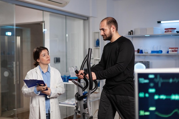 Hombre corredor en equipo deportivo trabajando en la resistencia del cuerpo durante un examen médico en un laboratorio moderno mientras que la investigadora analiza los datos de EKG que miden el pulso de la frecuencia cardíaca. Servicio de medicina