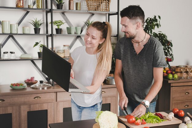 Hombre cortar verduras con cuchillo mirando a la mujer sosteniendo la computadora portátil en la cocina