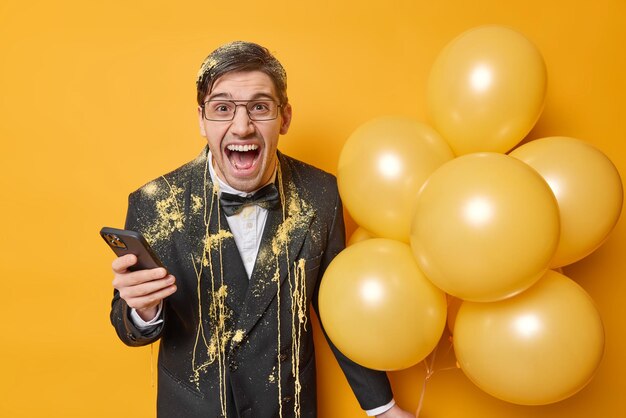 El hombre de cumpleaños enojado emocional grita en voz alta vestido con un elegante traje negro sostiene el teléfono móvil espera la llamada recibe poses de felicitación con globos inflados aislados sobre fondo amarillo