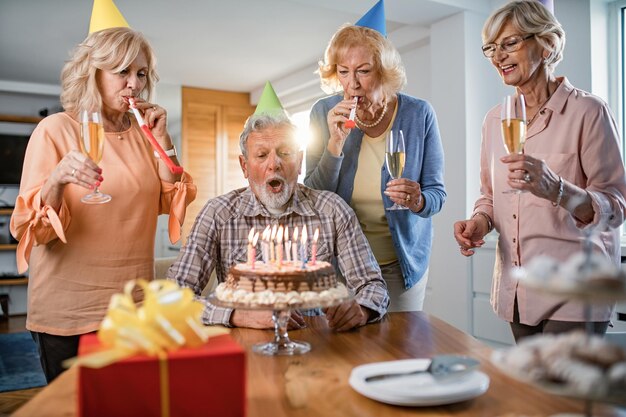 Hombre de cumpleaños maduro que sopla velas en el pastel mientras celebra el cumpleaños con sus amigos en casa