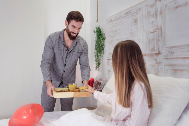 Hombre dando bandeja de madera con desayuno romántico a mujer.