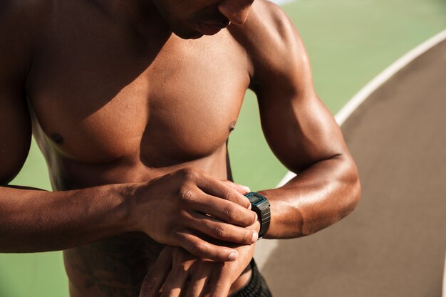 Hombre de deportes musculares afroamericanos sin camisa que controla el tiempo en el reloj de pulsera después del entrenamiento