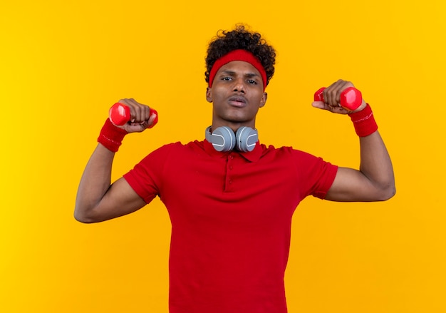 Hombre deportivo afroamericano joven confiado con diadema y muñequera con auriculares en el cuello levantando pesas aislado sobre fondo amarillo