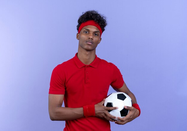 Hombre deportivo afroamericano joven confiado con diadema y muñequera sosteniendo la bola aislada en la pared azul